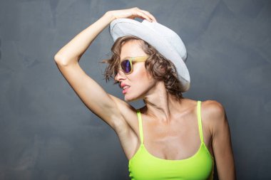 Beyaz şapkalı, güneş gözlüklü ve sarı mayolu gri arka planda poz veren genç modelin portresi.