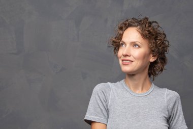 Gülümseyen esmer bir kadının portresi. Kıvırcık saçlı, gri arka planda günlük kıyafetler giyen.