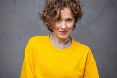 Kısa kıvırcık saçlı, parlak sarı süveter giyen, gri arka planda doğal makyajı olan mutlu, genç bir kadın.