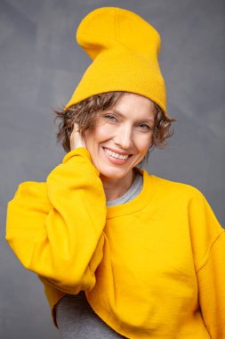 Sonbahar havası! Kısa esmer, kıvırcık saçlı, sarı kazaklı ve gri duvarlı arka planda şapkası olan mutlu bir kadının portresi.