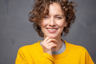 Kısa kıvırcık saçlı, parlak sarı süveter giyen, gri arka planda doğal makyajı olan mutlu, genç bir kadın.