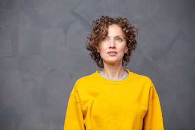 Kısa kıvırcık saçlı, parlak sarı süveter giyen, gri arka planda doğal makyajı olan mutlu, genç bir kadın.