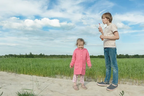Братья и сестры дети веселятся, делясь мыльными пузырями в зеленом летнем овсяном поле — стоковое фото