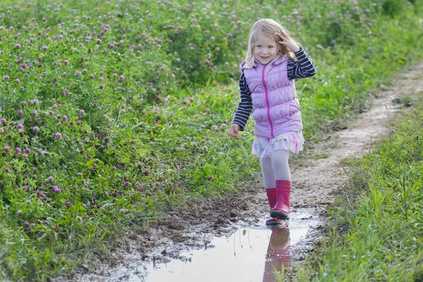 Uśmiechnięta blondynka z luźne jasne włosy, chodzenie na polnej drodze kałuży deszczu na łące kwiatów koniczyny fioletowy — Zdjęcie stockowe