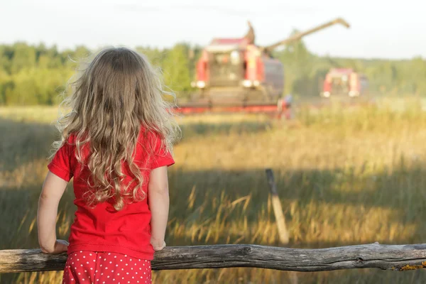 Chica rubia de granja en sartenes de lunares rojos para niños mirando el campo con cosechadora cosechadora —  Fotos de Stock