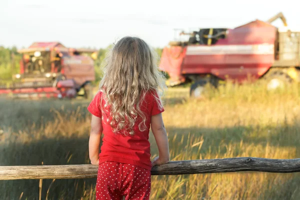 Pequeña chica de granja con sartenes de lunares rojos para niños mirando el campo con cosechadoras de trabajo —  Fotos de Stock