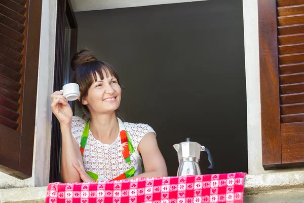 Porträtt av ung kvinna njuter av en kopp nybryggt kaffe med italiensk moka potten sitter nära öppna fönstret med traditionella Europeiska trä brun fönsterluckor — Stockfoto