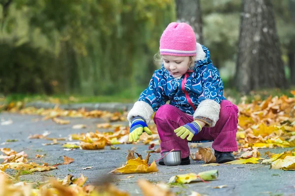 Dos años de edad, niña estirando su mano a metal termo frasco taza hunkering en otoño follaje cubierta telón de fondo — Foto de Stock