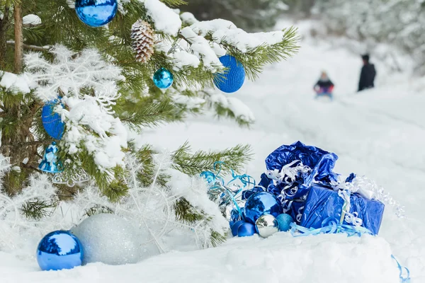 Рождественская снежная сосна украшена блестящими безделушками и двумя детьми, катающимися на санках по снежному покрову Стоковая Картинка