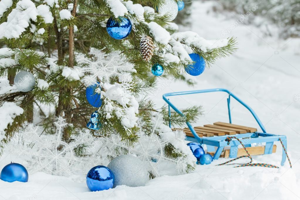 Christmas snowy pine tree decorated with glitter baubles and blue sledge on snow covering 