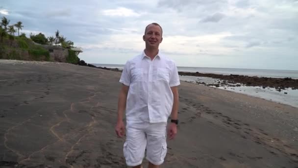 若い男が海を背景に歩いている 白いシャツとショートパンツを着た男が黒い砂浜を歩いている ロマンチックな朝の夜明け メディア — ストック動画