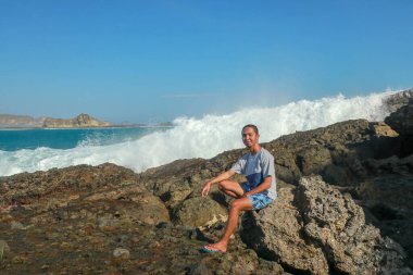 Tişörtlü ve mavi şortlu genç adam kayalık bir sahilde oturuyor. Bu deniz kıyısındaki kayanın üzerinde oturan bir adamın görüntüsü..