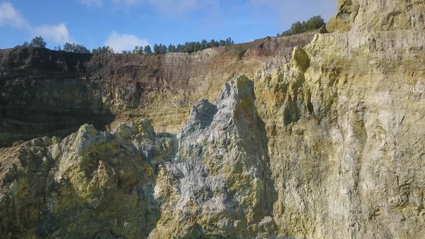 火山湖の上のクレーターのカラフルな壁への眺めを閉じます。ドローンは色付きの湖の表面の上に戻ってきます。太陽の光が周囲の岩を照らし — ストック写真