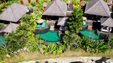 Tropikal ormanlarda ve palmiye ağaçlarında saman çatı villaları ve havuzları olan lüks otelin insansız hava aracı görüntüsü. Lüks villa, ormandaki pavyon, Ubud, Bali