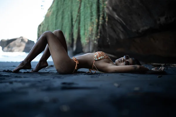 Belle fille au corps sexy en maillot de bain noir couché sur la plage avec du sable volcanique noir. Jeune modèle aux cheveux bouclés, peau bronzée posant et relaxant dans le voyage — Photo