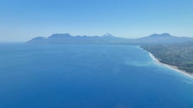 Captures a sweeping blue ocean meeting Flores volcanic coast, with distant mountains rising under a bright and clear sky.