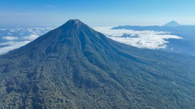 Volkanın güçlü konisi ufka hükmediyor. Etrafı sürüklenen bulutlar ve sonsuz ormanlarla çevrili. Endonezya doğasının vahşi özünü yakalıyor..