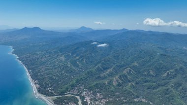 Verdant hills tumble toward the shoreline as distant volcanoes shape the horizon, painting a breathtaking panorama of untouched nature and expansive waters.
