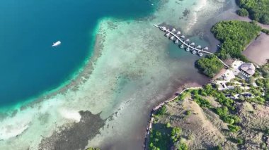 Yeşil tepelerle çerçevelenmiş mangrovlar ve açık deniz arasında yüzen su altı bungalovları ve Komodo adalarına giden botlarla dolu bir kanal..