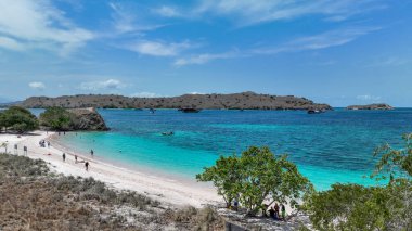Pastel kıyı şeridinin berrak suyla buluştuğu Komodo Pembe Plajı 'nın geniş bir manzarası ve vahşi, güneşli tepeler arasında yüzen sessiz tekneler..