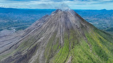 Tarlalar ve çiftlikler yanardağın eteklerinde huzur içinde uzanır. Bereketli yeşil toprak ile kara kara düşünen, dumanlı tepe noktası arasında canlı bir tezat vardır..