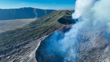 Bromo aktif külahının destansı manzarası sisli vadiler ve yeşil tepeler arasında beyaz tüyler püskürüyor, doğanın büyüleyici ateşli nefesi.