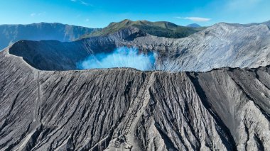 İz, Bromo 'nun dumanı tüten mavi kalbine kadar uzanır. Dalgalı kül yamaçları ve uzak sisli tepelerle çevrilidir..