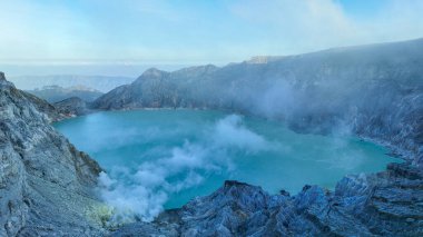 Kawah İjen 'in puslu uçurumları turkuaz krater gölü üzerinde yükseliyor, uzak volkanik tepeler yumuşak mavi sabah sisine dönüşüyor..