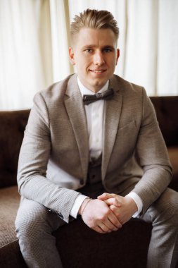 Bir adamın portresi. Gri takım elbiseli, beyaz gömlekli ve papyonlu damat kahverengi deri koltukta oturuyor. Koltukta oturan takım elbiseli damat. Yüksek kalite fotoğraf