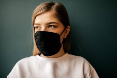 Siyah maskeli genç bir kadının portresi koyu yeşil arka planda izole edilmiş. Genç kız hasta duvarın arkasında duruyor, covid-19, Coronavirus 'u durdur..