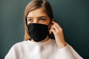 Siyah maskeli genç bir kadının portresi koyu yeşil arka planda izole edilmiş. Genç kız hasta duvarın arkasında duruyor, covid-19, Coronavirus 'u durdur..