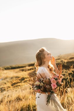 Beyaz gelinlikli güzel gelin, bir buketle dağlarda poz veriyor ve gülümsüyor. Yüksek kalite fotoğraf