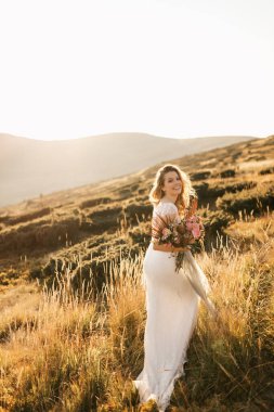 Beyaz gelinlikli güzel gelin, bir buketle dağlarda poz veriyor ve gülümsüyor. Yüksek kalite fotoğraf