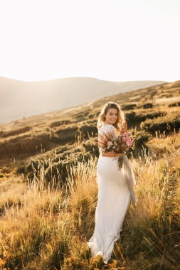 Beyaz gelinlikli güzel gelin, bir buketle dağlarda poz veriyor ve gülümsüyor. Yüksek kalite fotoğraf