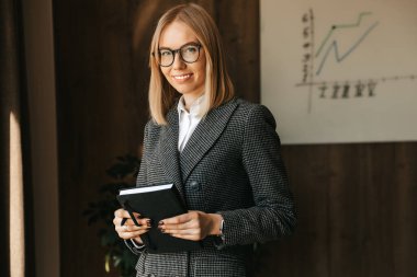 Bir iş kadını elinde bir defter tutuyor ve şirketine yeni projeler planlarken ofiste gülümsüyor.