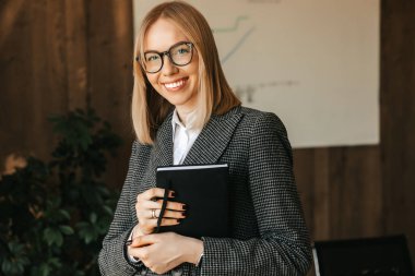 Gözlüklü, takım elbiseli ve tişörtlü güzel bir beyaz kadın çalışma masasının yanında duruyor elinde bir defter, ve ofiste dişlek bir gülümsemeyle gülümsüyor..