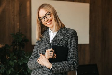 Gözlüklü, takım elbiseli ve tişörtlü güzel bir beyaz kadın çalışma masasının yanında duruyor elinde bir defter, ve ofiste dişlek bir gülümsemeyle gülümsüyor..