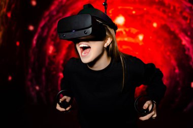 Duygusal olarak genç bir kız sanal gerçeklik gözlükleriyle bir oyun oynuyor. Elinde av tüfekleri var. Oyun odasında parlak kırmızı arka plan. Yüksek kalite fotoğraf