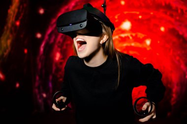 Duygusal olarak genç bir kız sanal gerçeklik gözlükleriyle bir oyun oynuyor. Elinde av tüfekleri var. Oyun odasında parlak kırmızı arka plan. Yüksek kalite fotoğraf