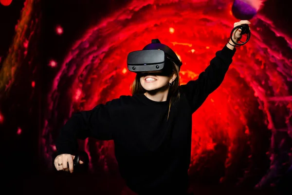 The girl emotionally plays the game in virtual reality glasses holding gamepads in her hands. Red game background. High quality photo