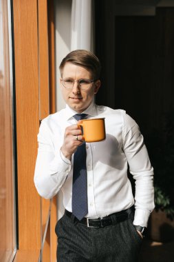 Genç işadamı elinde bir fincan kahveyle ofisinin penceresinde duruyor..