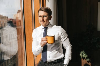 Genç işadamı elinde bir fincan kahveyle ofisinin penceresinde duruyor..