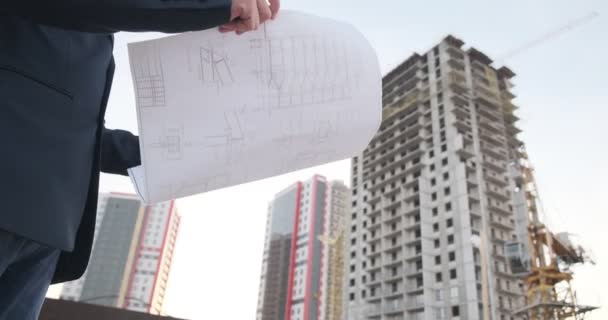 젊은 남자 건축가가 건물의 건축을 조사 한다. 한 기술자가 길가에 있는 건물의 건설 현장과 반대되는 그림을 보고 있습니다. — 비디오