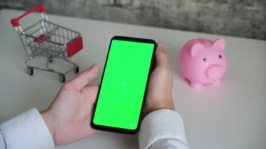 Yakın plan çekimde kromakey ekranlı bir telefon. Üst görünüm akıllı telefon kumbarası ve alışveriş arabası beyaz arkaplanda kopyalama alanı