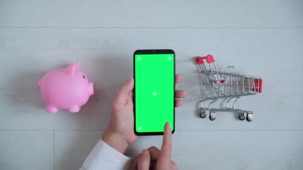 Telefone com uma tela chromakey nas mãos de um close-up. top view smartphone piggy Bank e carrinho de compras espaço de cópia no fundo branco — Vídeo de Stock