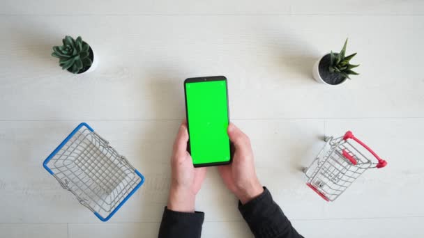 Een telefoon met een groen chromakey scherm en een winkelwagentje op een witte achtergrond. bovenaanzicht van de smartphone in de handen van een close-up — Stockvideo
