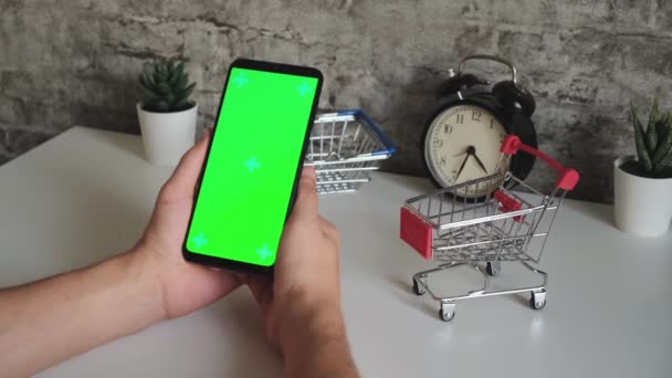 Mesa con cestas de compras primer plano teléfono móvil en las manos con pantalla verde. pasa en la pantalla cromakey teléfonos inteligentes en primer plano. vídeo listo para insertar y teclear contenido en línea — Vídeos de Stock