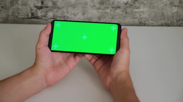 Un hombre en un escritorio blanco está sentado con un teléfono móvil en sus manos con una pantalla verde. deslizamientos y grifos en la pantalla cromakey teléfonos inteligentes en primer plano. Vídeo listo para insertar y teclear — Vídeo de stock