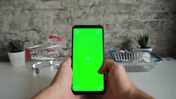 Alışveriş sepetleri olan bir masa. Elinde yeşil ekranlı cep telefonu var. Akıllı telefonların kromakey ekranını yakın planda tutuyor. İçeriği çevrimiçi yerleştirmek ve anahtarlamak için hazırlanmış video — Stok video