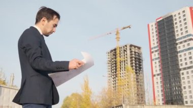 Genç bir erkek mimar binanın inşaatını denetliyor. Bir mühendis, binanın arka planındaki çizimlere bakıyor.